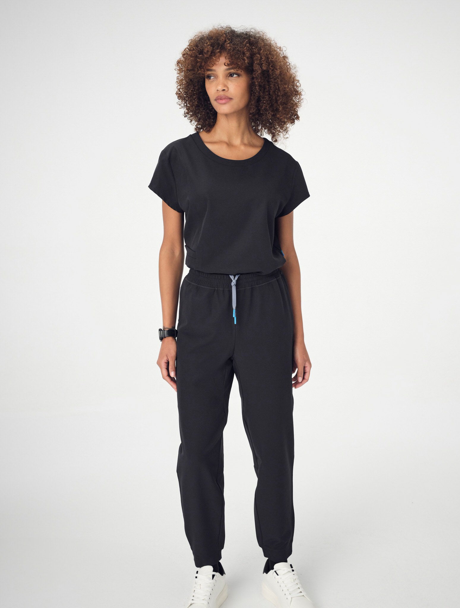 A woman posing in a set of black Penna scrubs and white running shoes.