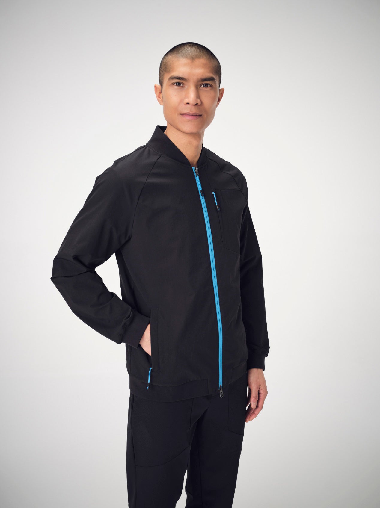 A man posing with his right hand on his hip, wearing black Penna scrubs. He is wearing the Falcon jogger pants and a Bomber style jacket.
