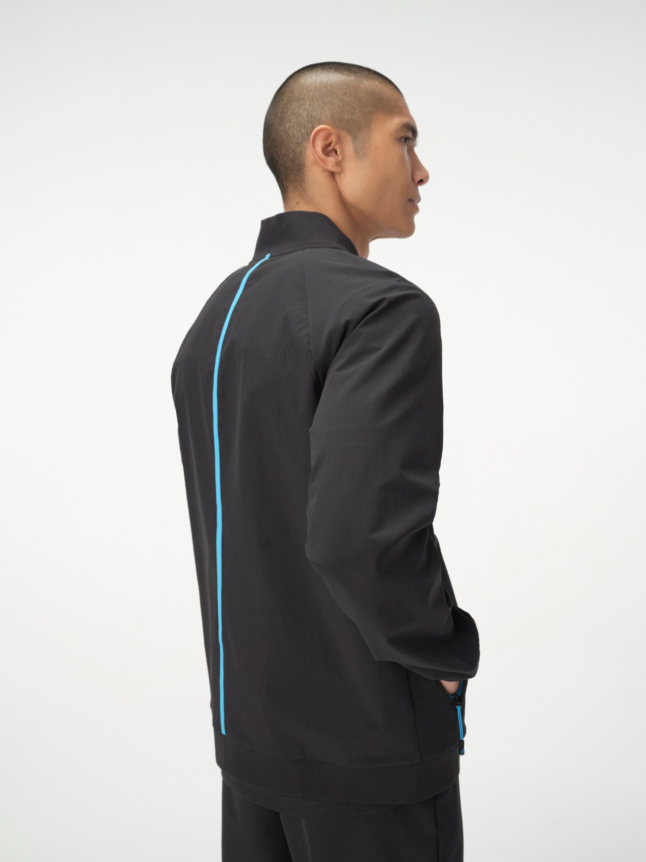 A man photographed from behind wearing a set of black Penna scrubs. He is wearing the bomber-style jacket, The Eagle, which has a bright blue front zipper and a bright blue seam down the length of the back and the Falcon Jogger pants.