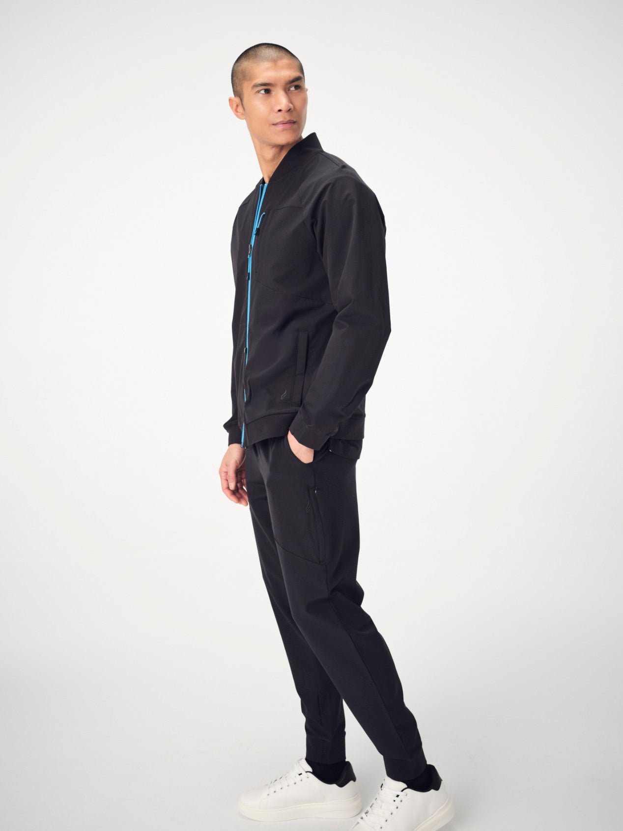 A man photographed from behind wearing a set of black Penna scrubs. He is wearing the bomber-style jacket, The Eagle, which has a bright blue front zipper and a bright blue seam down the length of the back and the Falcon Jogger pants.