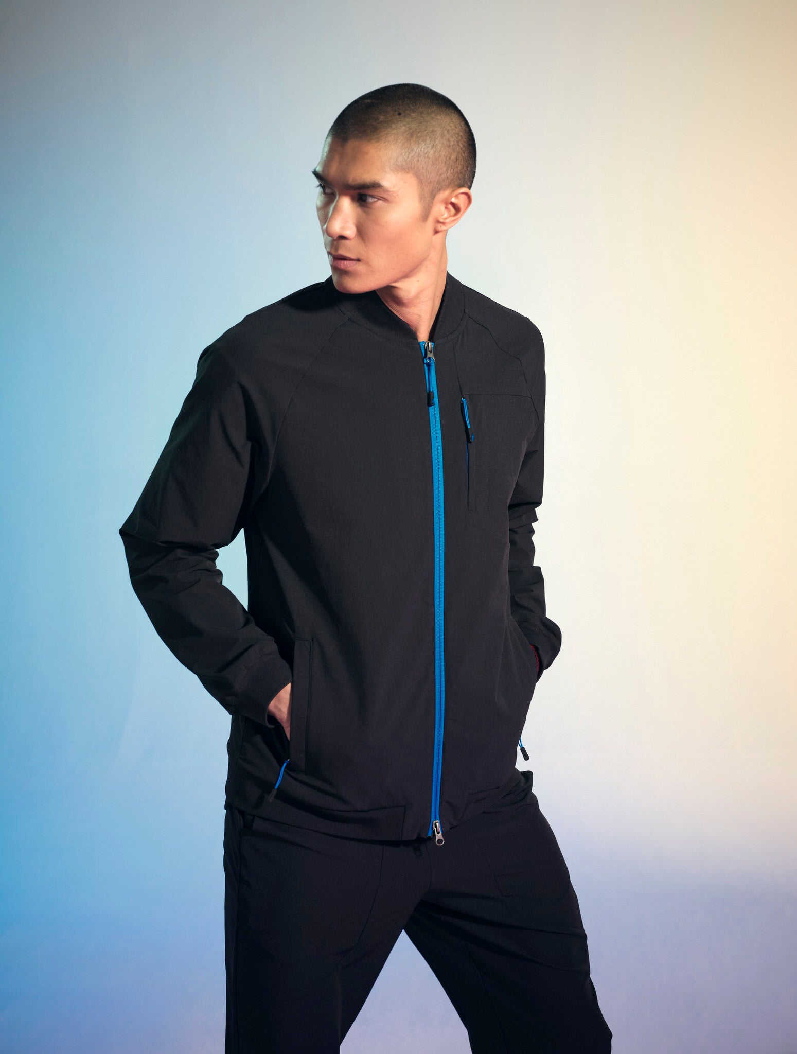 A man wearing a set of black Penna scrubs posing with his hands in his pockets and looking off to the left. He is wearing the The Eagle bomber jacket, which has a bright blue front zipper and a bright blue seam down the back and the Falcon Jogger pants.