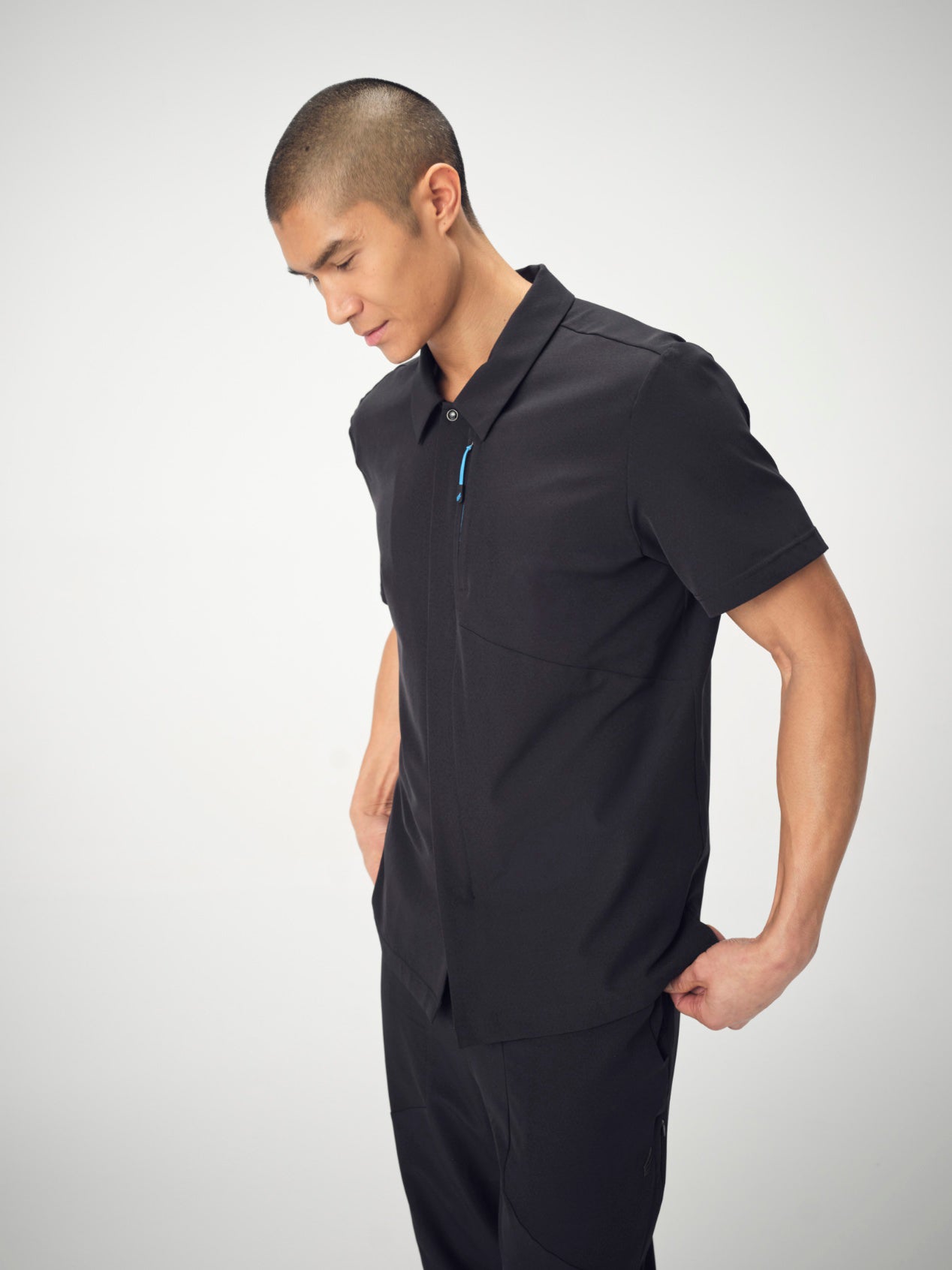 A man photographed from the knees up is wearing black Penna scrubs. He is looking down and posed with his hands in his pockets.