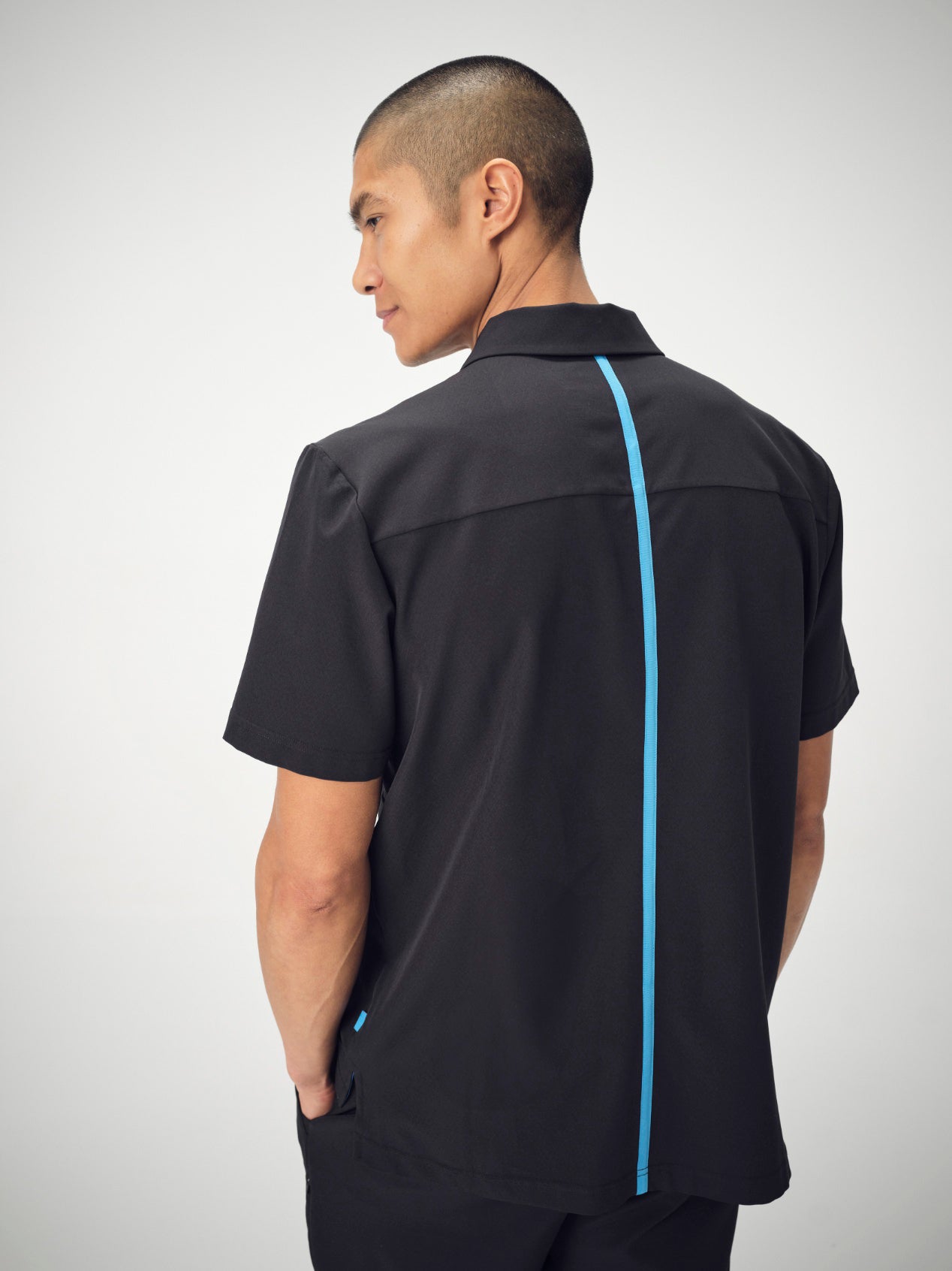 A man photographed from behind, from the waist-up, is wearing a black Penna Men's short-sleeved collared shirt style The Hawk. He is posed with his hands in his pockets and is looking to the left.