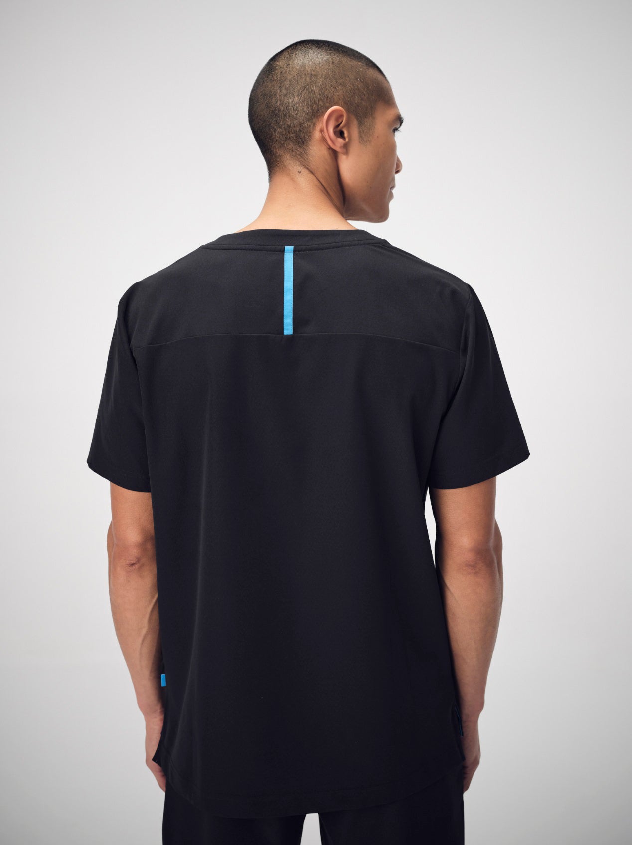 A man photographed from behind wearing a set of black Penna scrubs. He is wearing the Hawk, a short-sleeved style with a bright blue seam down the back yoke.