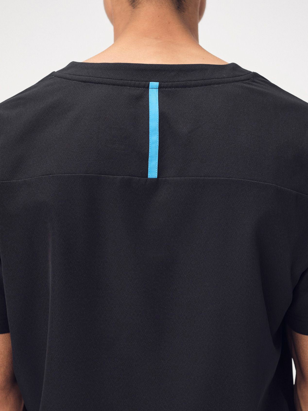 A closeup of a man's shoulders wearing the black Penna scrub top style, The Hawk, which is short-sleeved and has a bright blue seam on the back yoke.