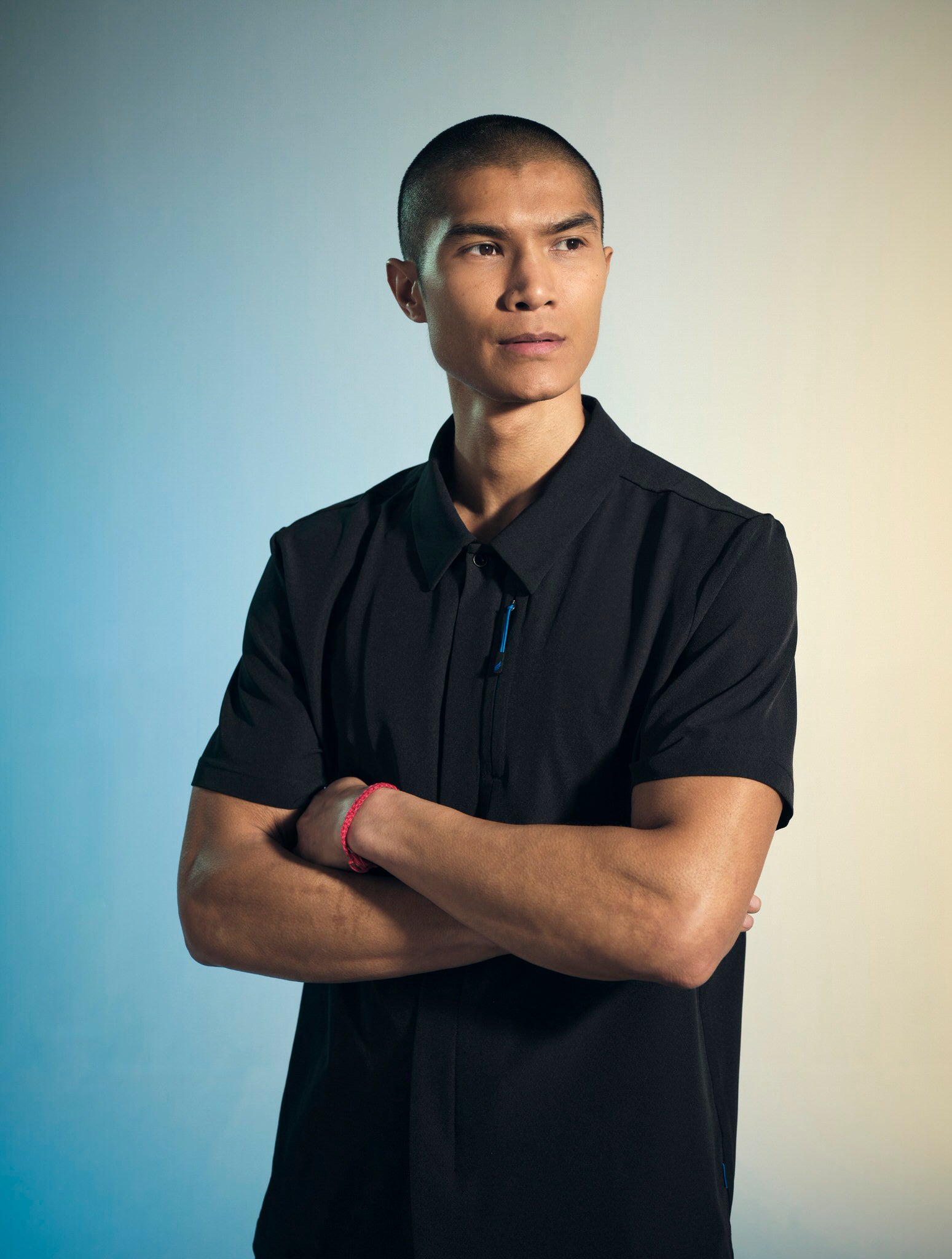 A man photographed from the waist-up, with a serious expression and his arms crossed, is wearing a black Penna, short-sleeved, collared shirt style The Hawk.