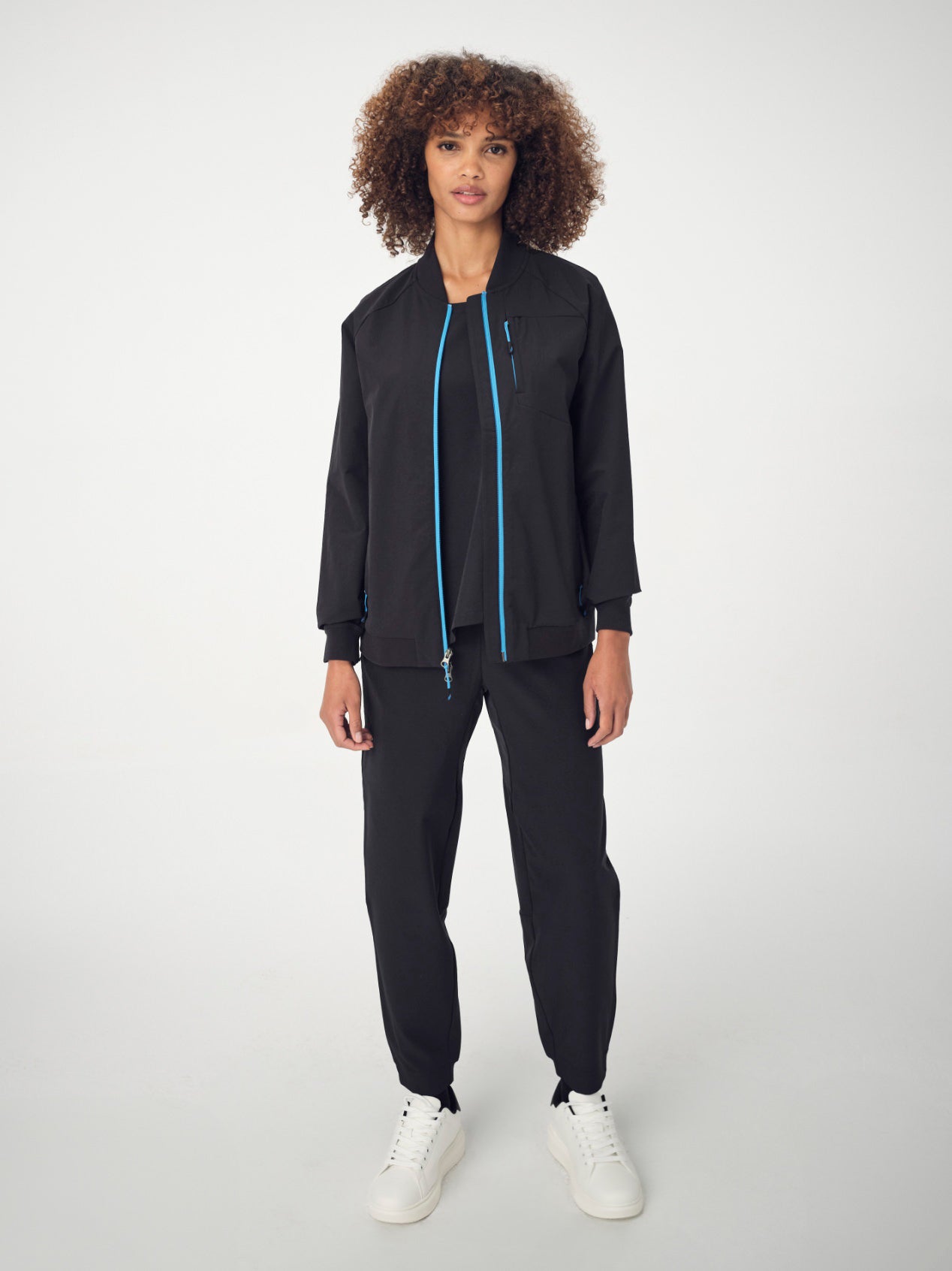 A woman with brown, curly, shoulder-length hair is wearing the black Penna bomber jacket style The Hummingbird, the Dove jogger bottoms, and white sneakers. She is facing the camera with her arms by her sides.