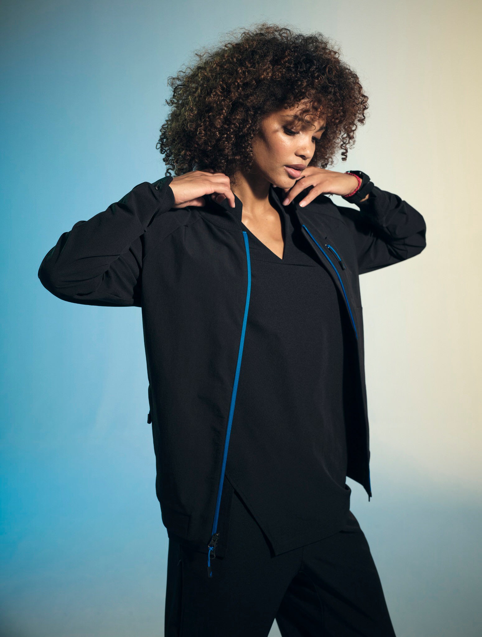 A woman with brown, curly, shoulder-length hair is adjusting the collar of her unzipped, black bomber jacket in The Hummingbird style. She is also a wearing black, Skylark V-neck short-sleeved top and the Dove jogger bottoms.