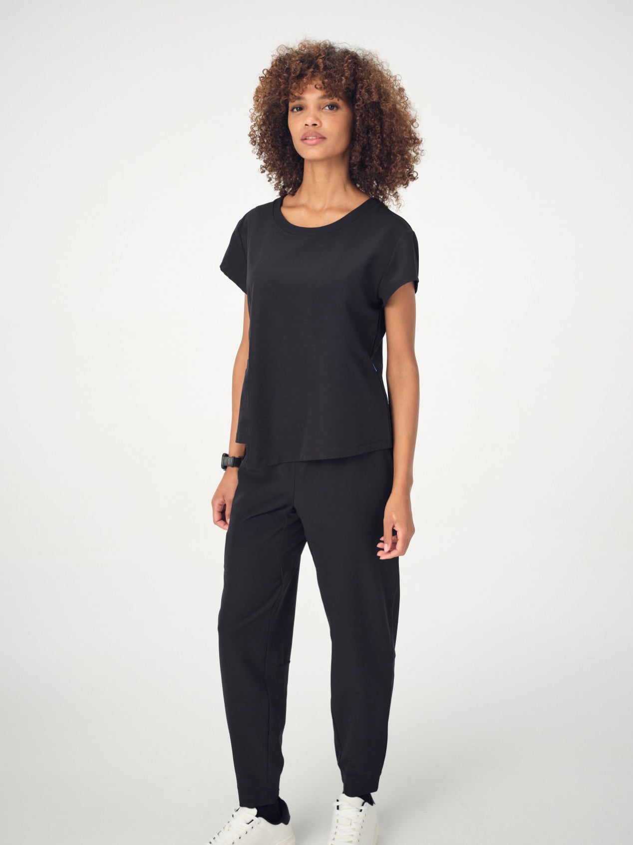 A woman with brown, curly, shoulder-length hair is wearing the black Penna Skylark crew-neck, short-sleeved top, The Dove jogger bottoms, and white sneakers. She is facing the camera with her arms by her sides.