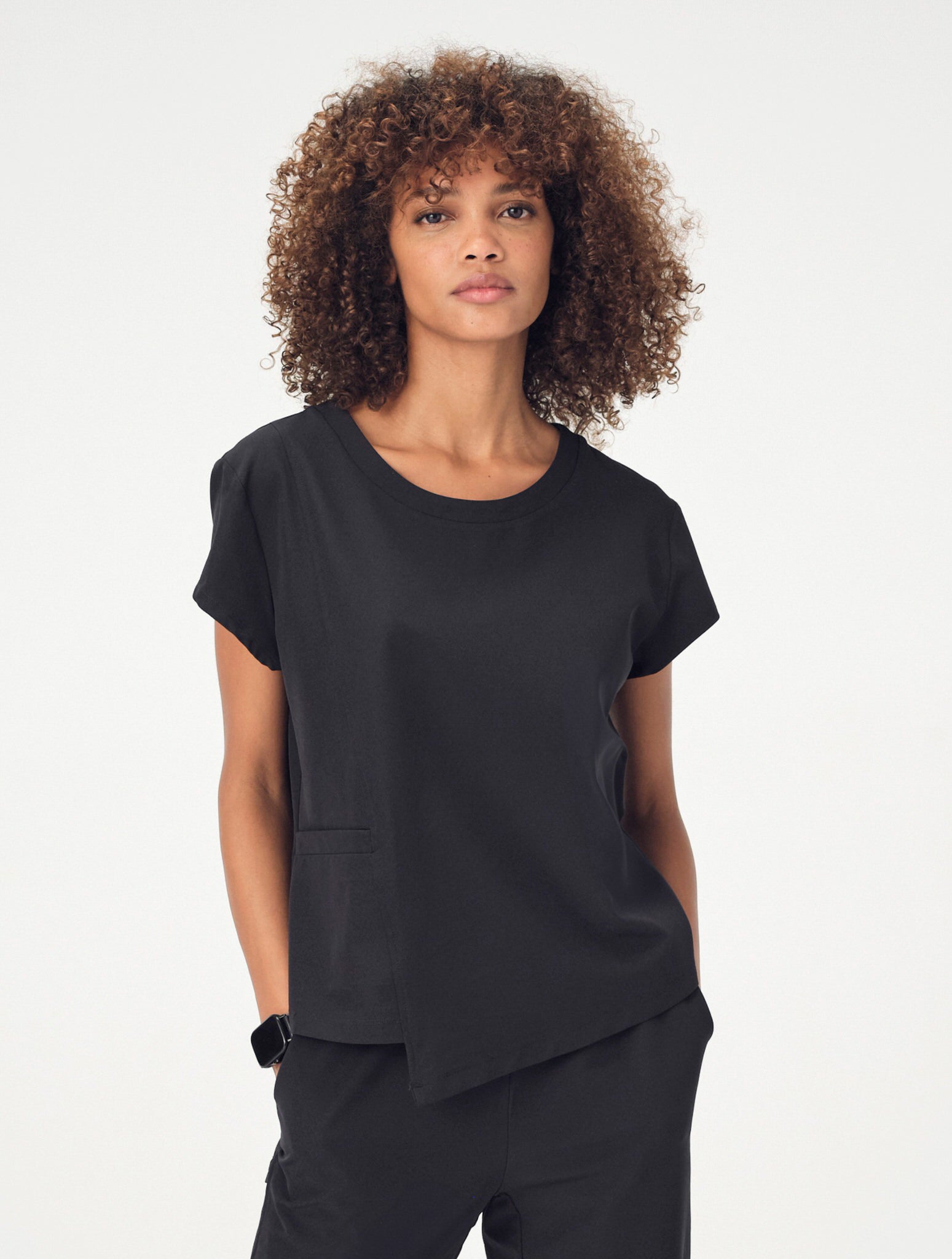A woman with brown, curly, shoulder-length hair is wearing the black Penna Skylark crew-neck, short-sleeved top, the Dove jogger bottoms, and white sneakers. She is facing the camera with her arms by her sides.