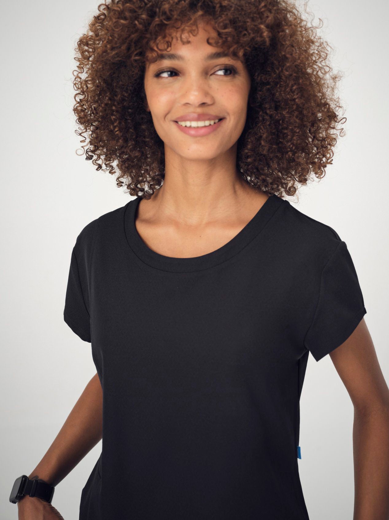 A woman with brown, curly, shoulder-length hair is wearing the black Penna Skylark crew-neck, short-sleeved top. She is smiling and looking to the left of the camera.