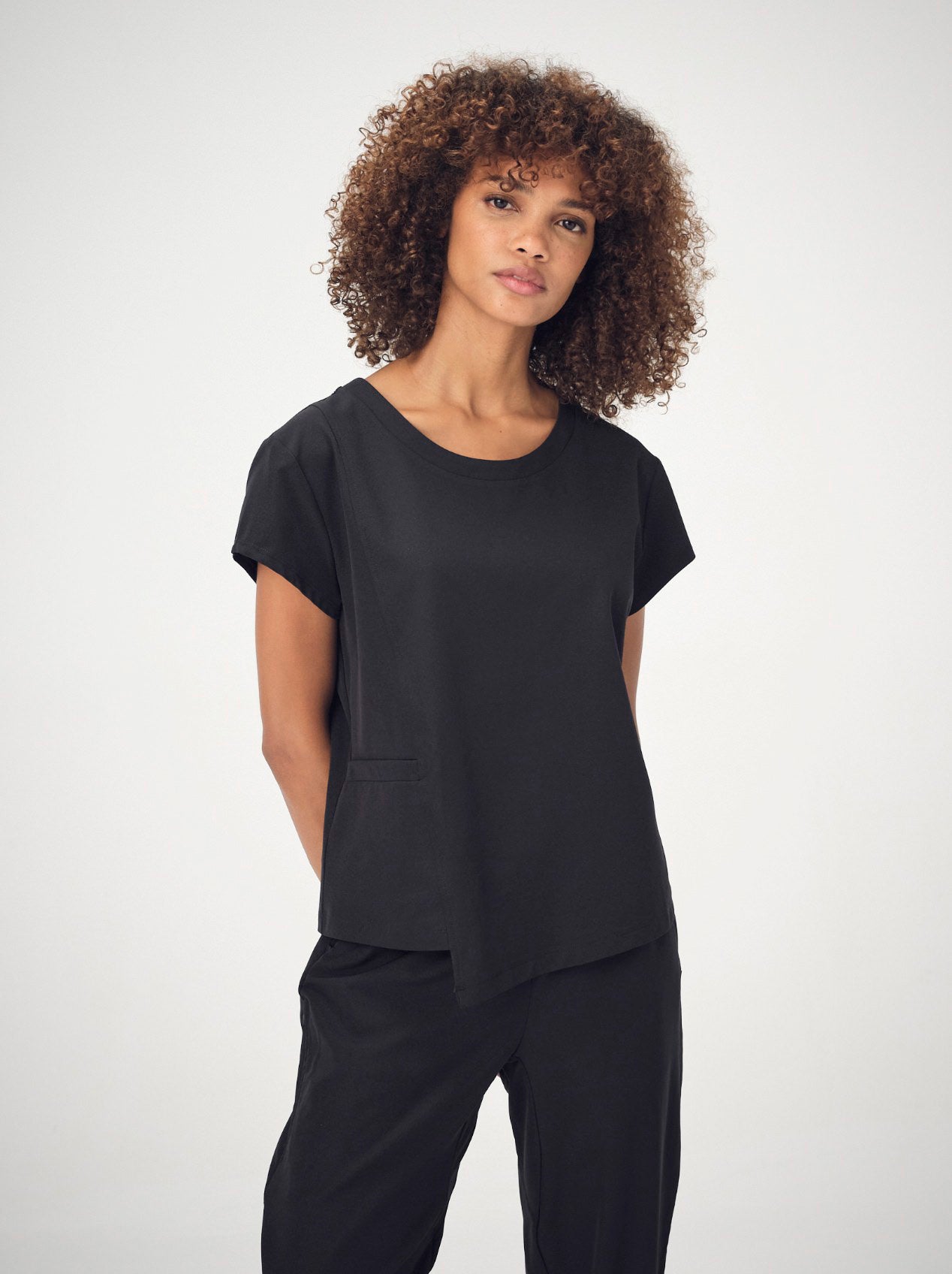 A woman with brown, curly, shoulder-length hair is wearing the black Penna Skylark crew-neck, short-sleeved top and the Dove jogger bottoms She is facing the camera with her arms behind her back.