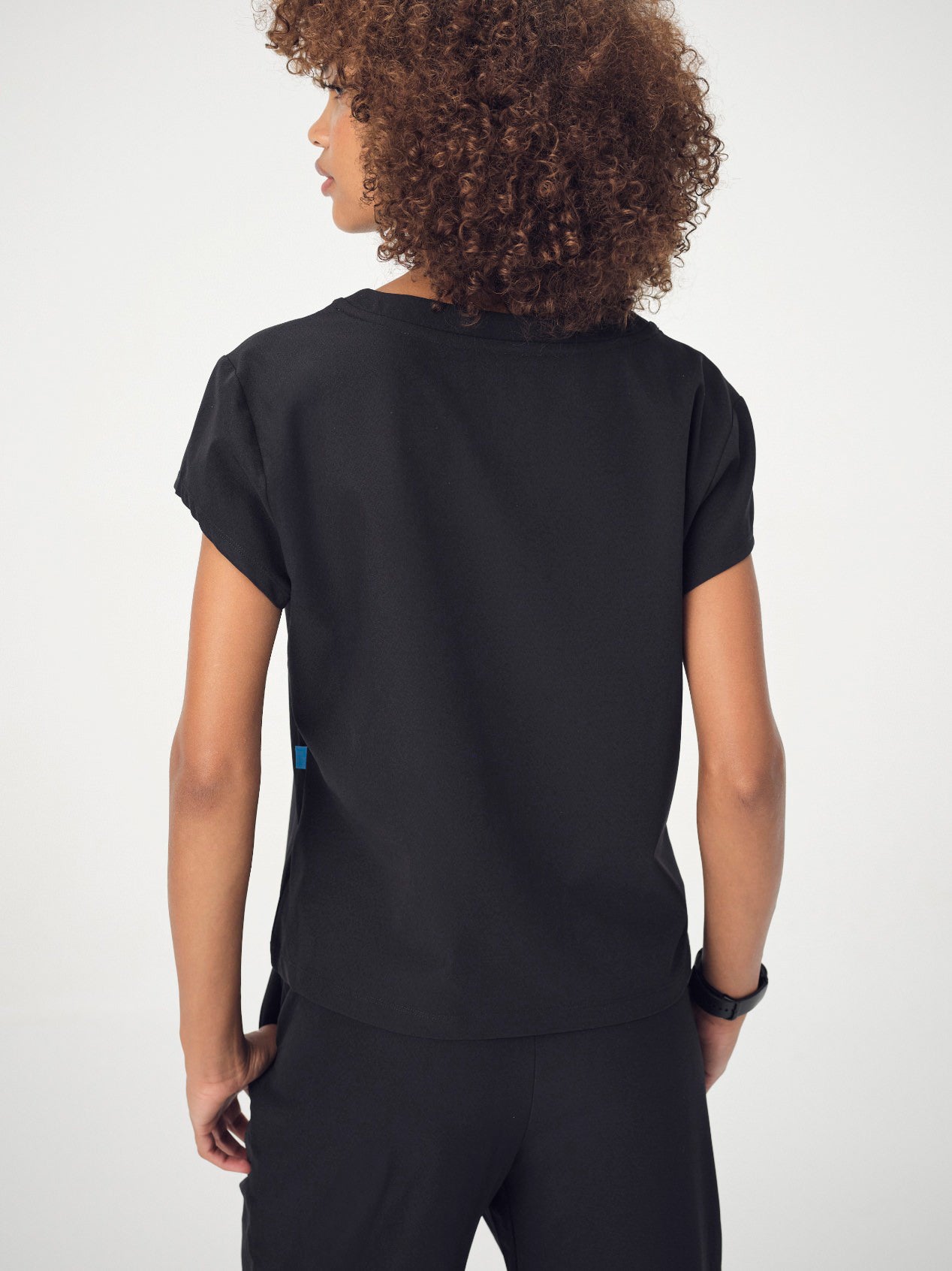 A woman with brown, curly, shoulder-length hair photographed from behind is wearing the black Penna Skylark crew neck, short-sleeved top and Dove jogger bottoms. She is posing with her hands in her pockets and looking to the left.