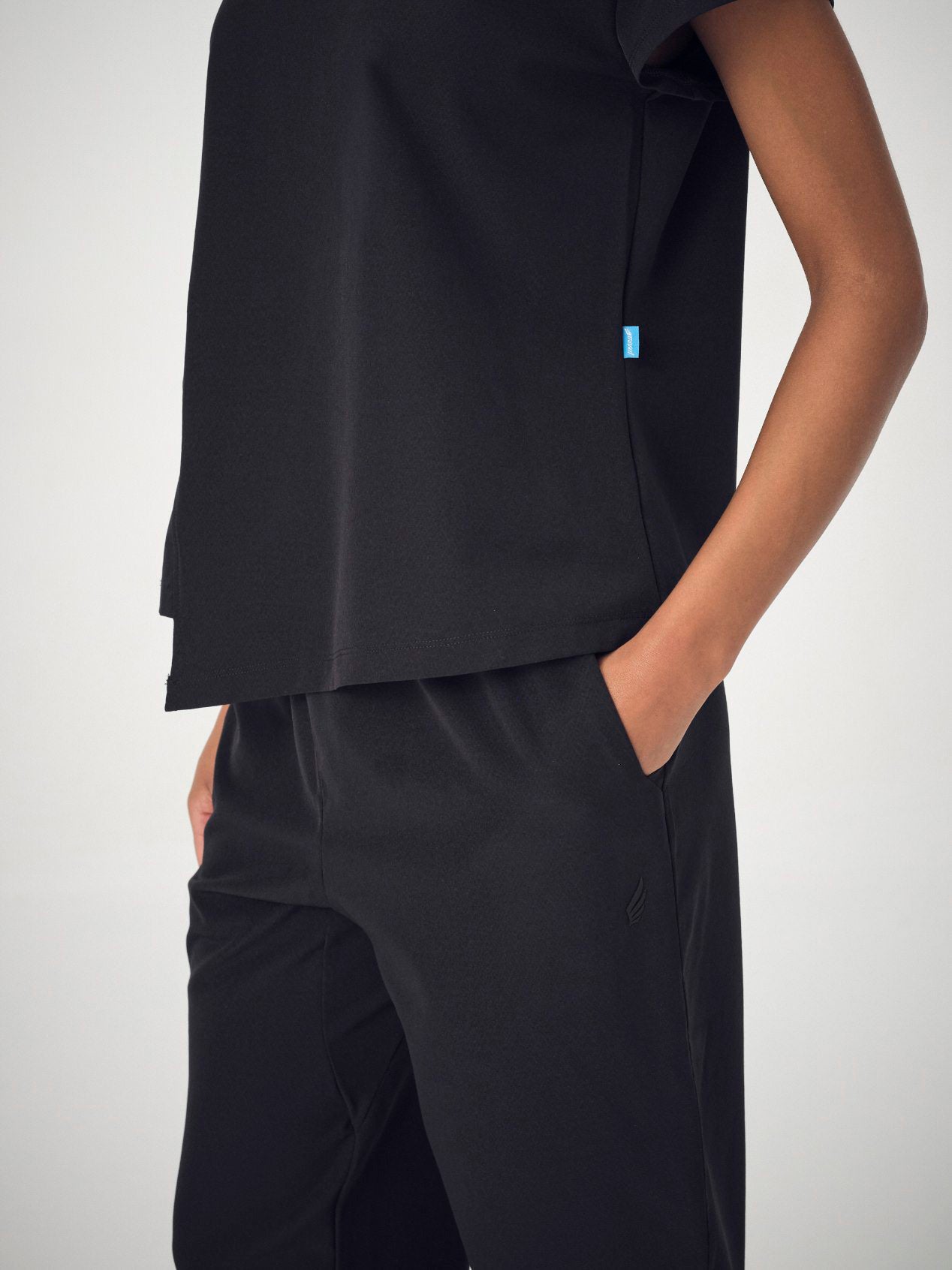A close-up of a woman's torso wearing the black Penna Skylark crew neck, short-sleeved top and the Dove jogger bottoms in black. The woman's hands are in her pants pockets.