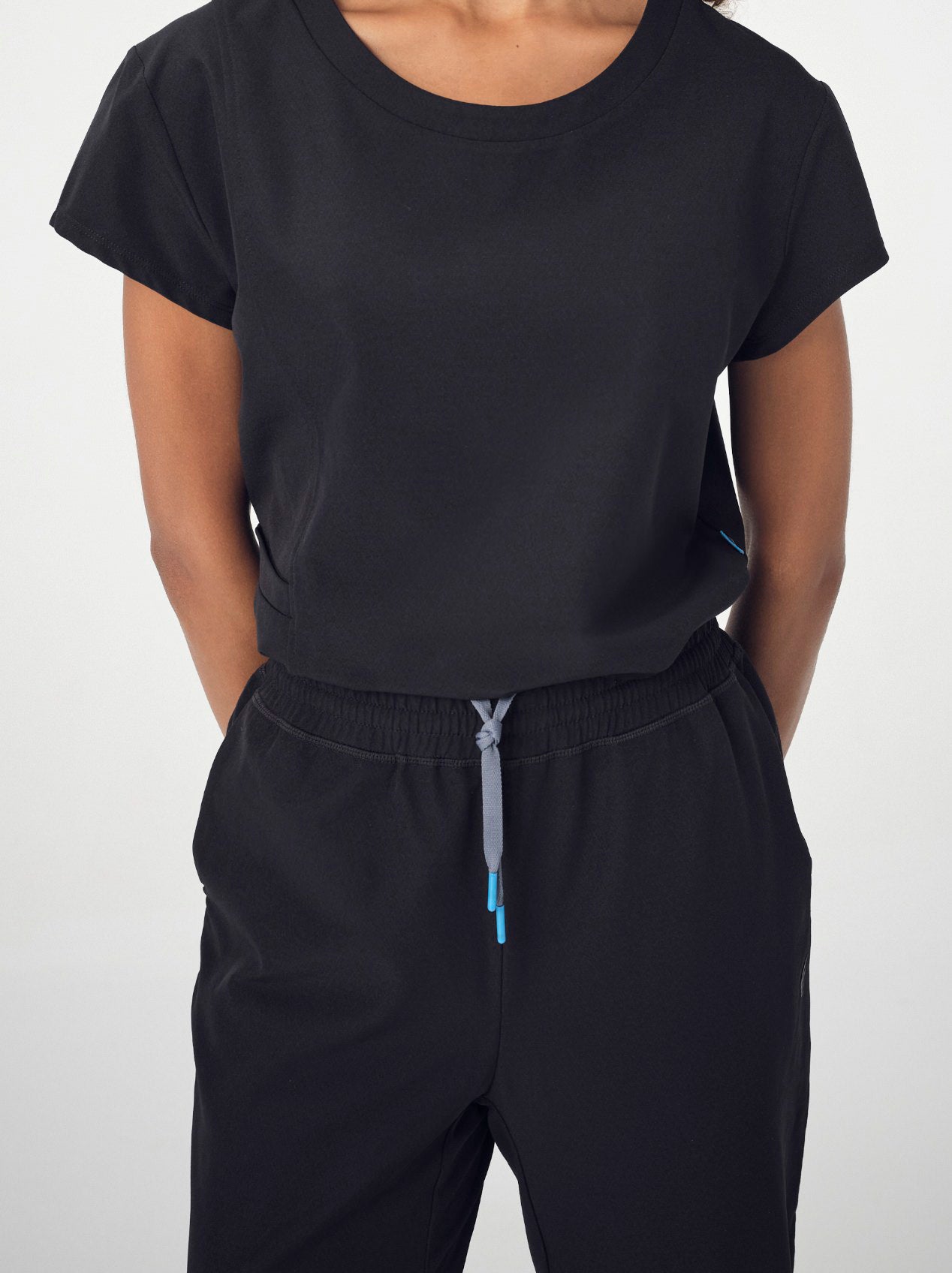 A close-up of a woman's torso wearing the black Penna Skylark crew neck, short-sleeved top and the Dove jogger bottoms in black. The woman's hands are clasped loosely behind her back.