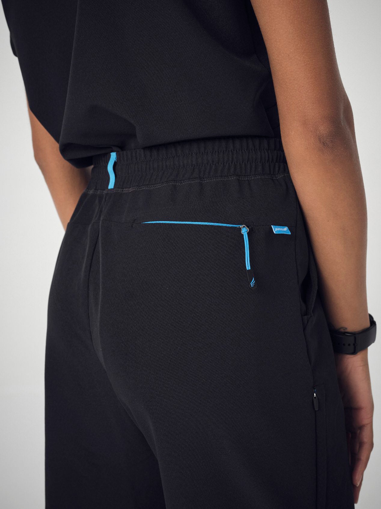 A closeup of from behind of a woman wearing a set of black Penna scrubs with the shirt tucked into the pants. She is wearing a black watch on her right wrist and her hands are by her sides.