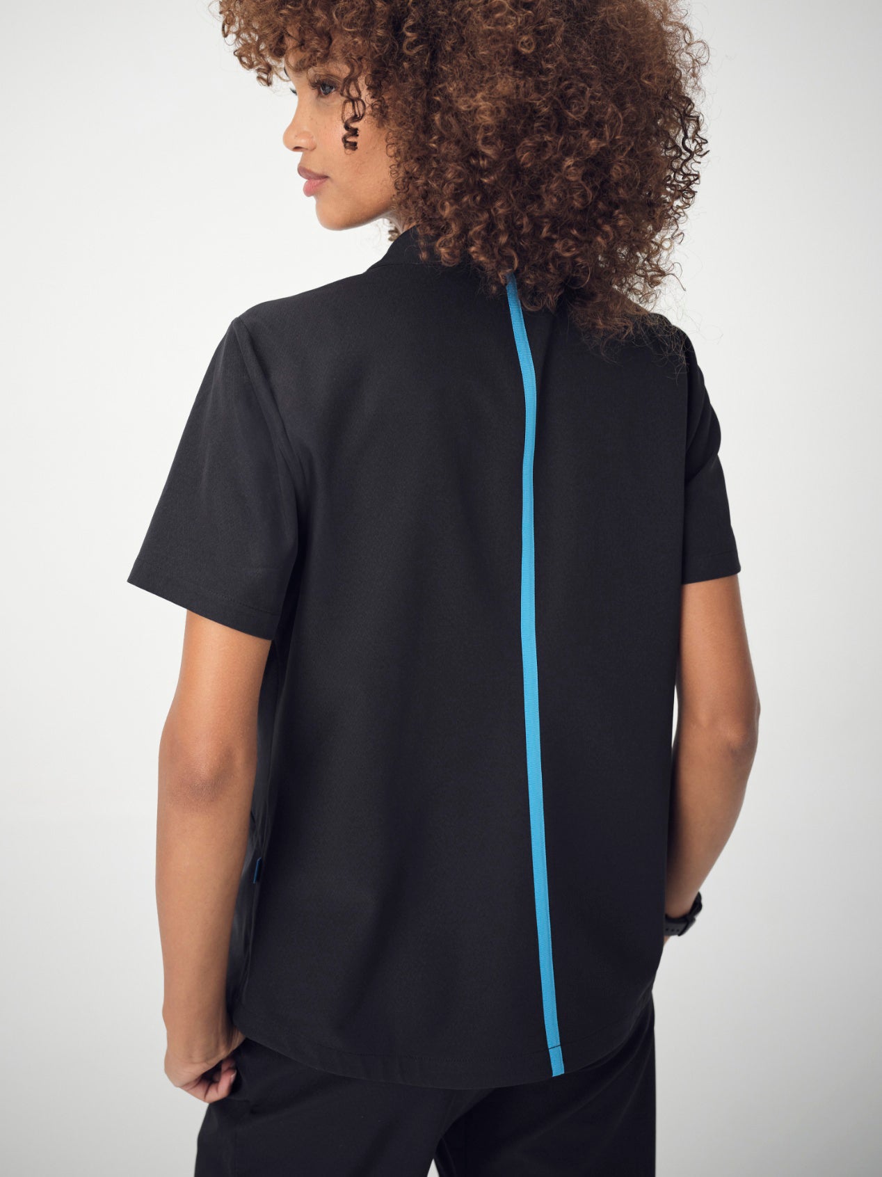 A woman with brown, curly, shoulder-length hair photographed from behind is wearing the black Penna Skylark pocket, short-sleeved top and Dove jogger bottoms. She is posing with her hands in her pockets and looking to the left.