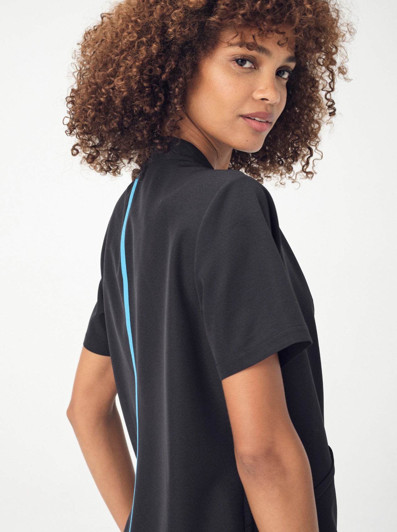 A woman with brown, curly, shoulder-length hair photographed from the waist up wearing the black Penna Skylark pocket, short-sleeved top. She is looking back over her right should into the camera.