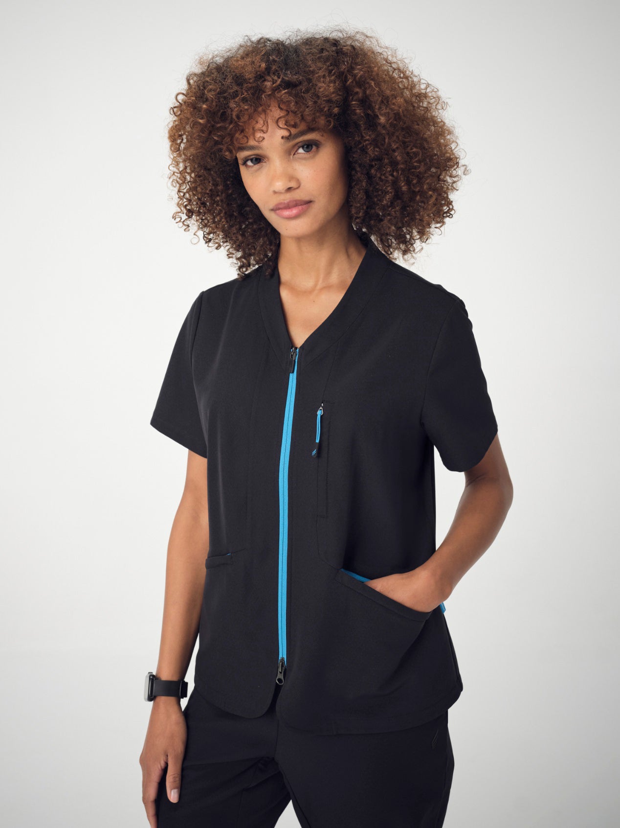 A woman photographed from the waist up, posing with her left hand in her pocket, wearing the black Penna Skylark short-sleeved, pocket top.
