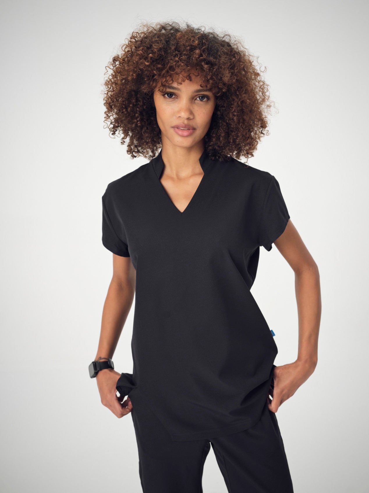 A woman photographed from the front, posing with her hands in her pockets, wearing the black Penna Skylark short-sleeved, v-neck top and black jogger bottoms in the Dove style.