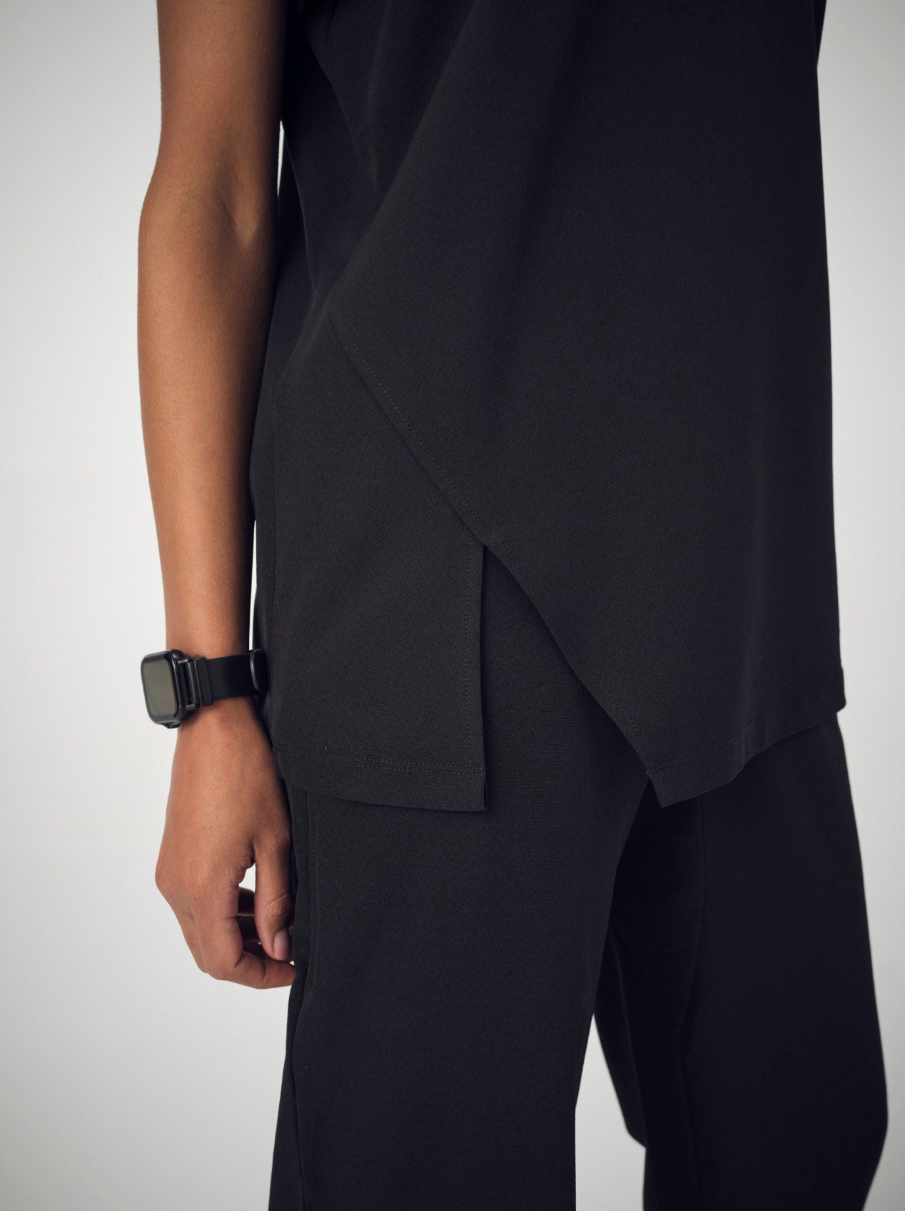 A closeup of the front of a woman wearing a set of black Penna scrubs with the shirt tucked into the pants. She is wearing a black watch on her right wrist.