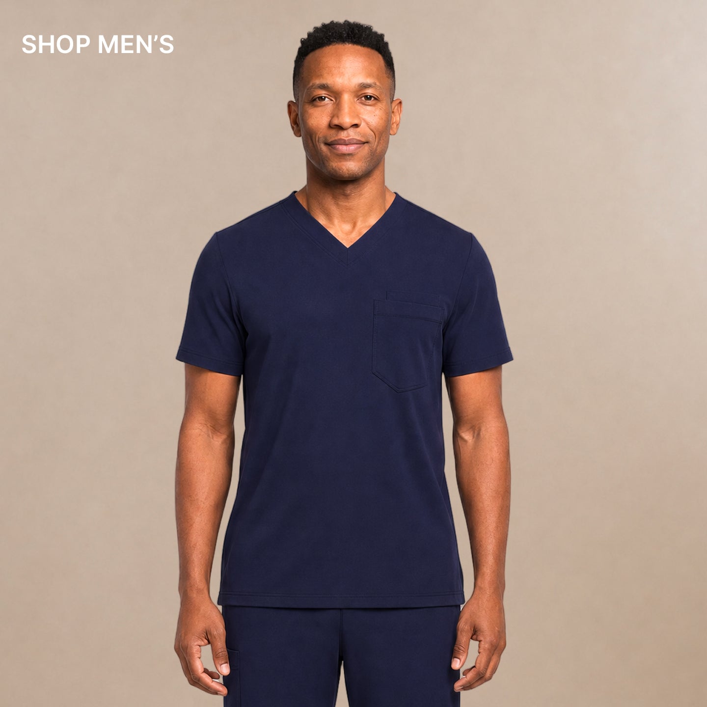 A man modeling a navy blue V-neck scrub top and matching scrub pants set, standing against a neutral beige studio background.