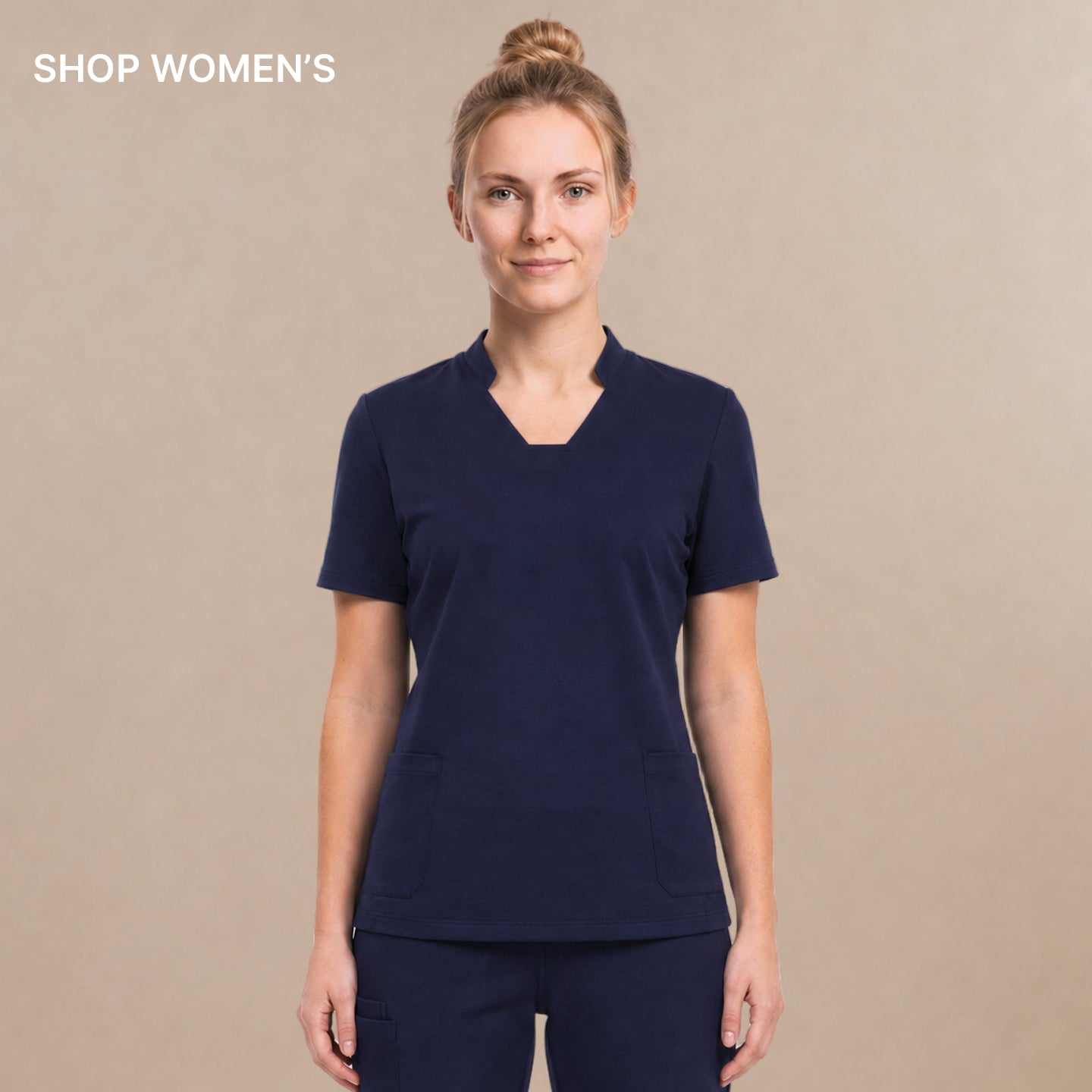 A woman modeling a matching navy blue scrub set consisting of a short-sleeve V-neck top and straight-leg pants, standing against a solid beige studio background.