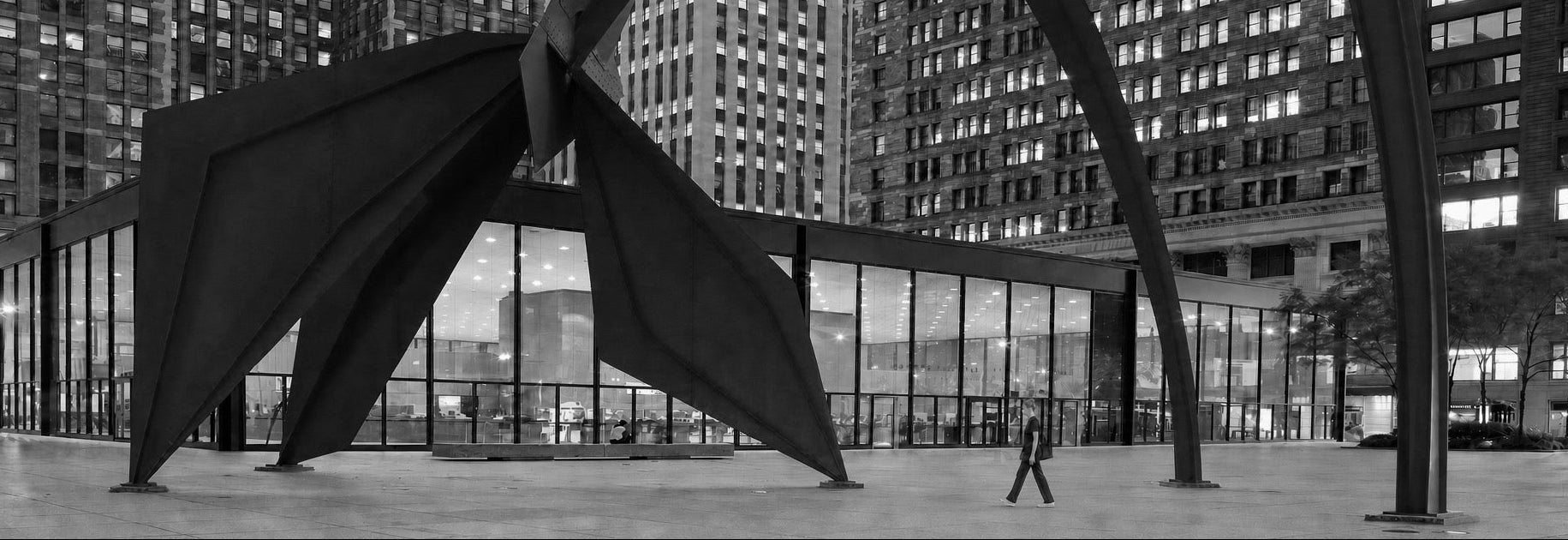 Black and white photograph of a large abstract metal sculpture in an urban plaza in Chicago, with tall buildings in the background and a woman wearing scrubs walking by.