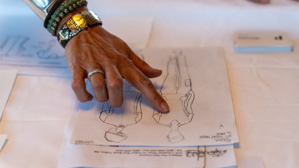 A close-up of a person's hand with rings and bracelets pointing to a hand-drawn fashion sketch on paper, set on a light-colored table.