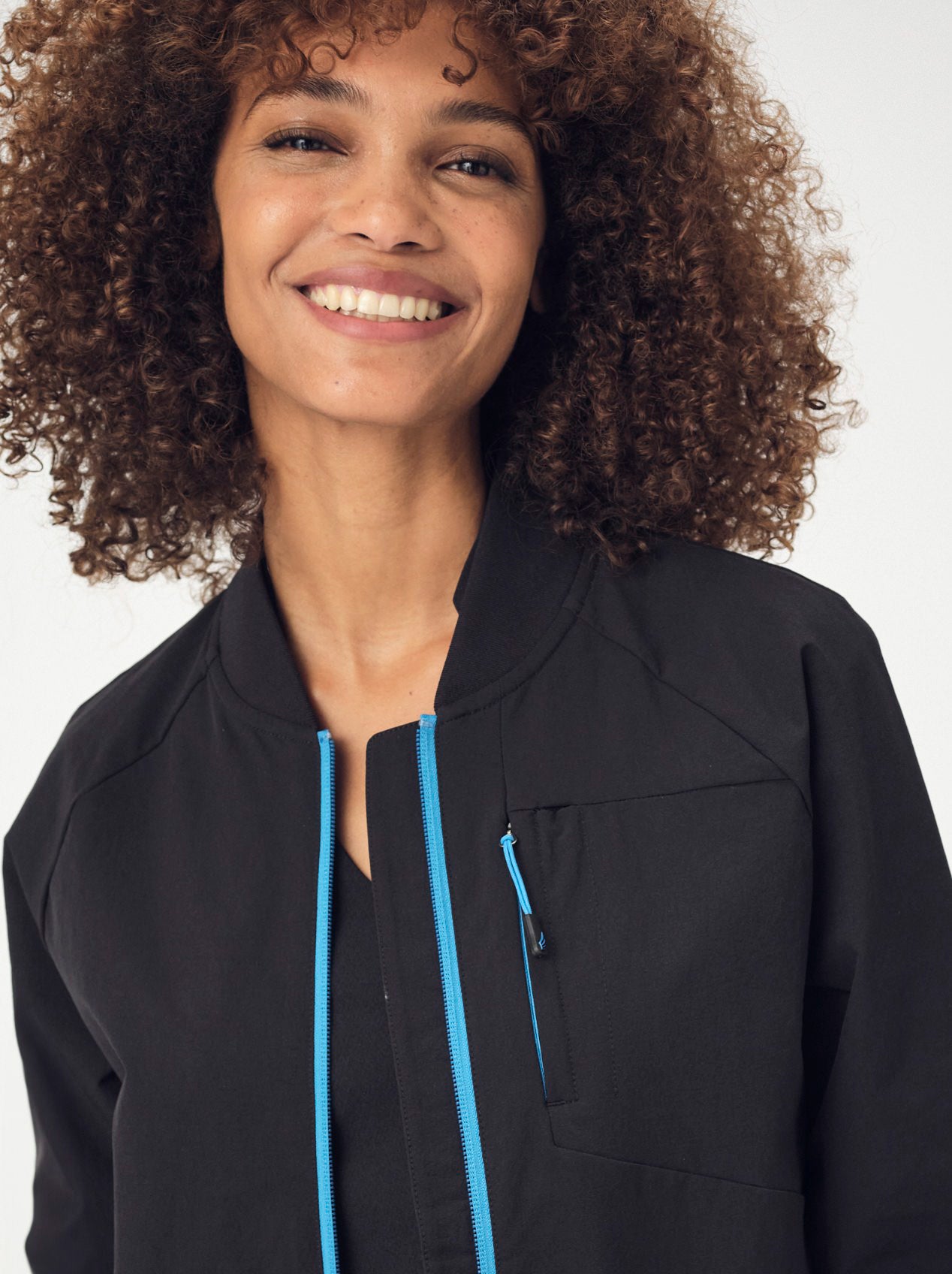 A smiling woman with brown, curly, shoulder-length hair is photographed from the chest-up wearing an unzipped black Penna bomber jacket The Hummingbird and a black top underneath.