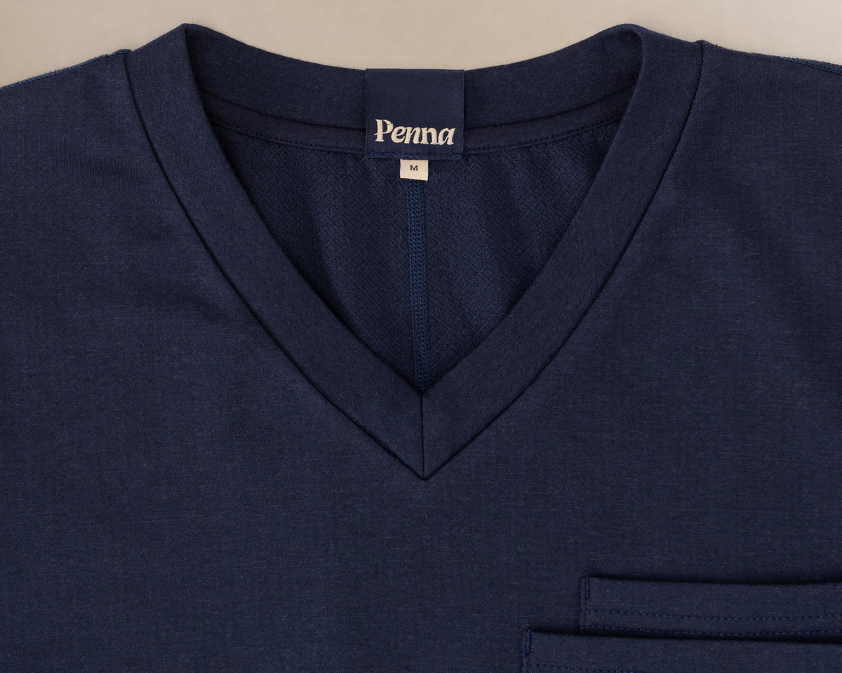 Close-up of a navy blue V-neck scrub t-shirt with a left chest pocket and a visible 'Penna' brand label inside the collar.