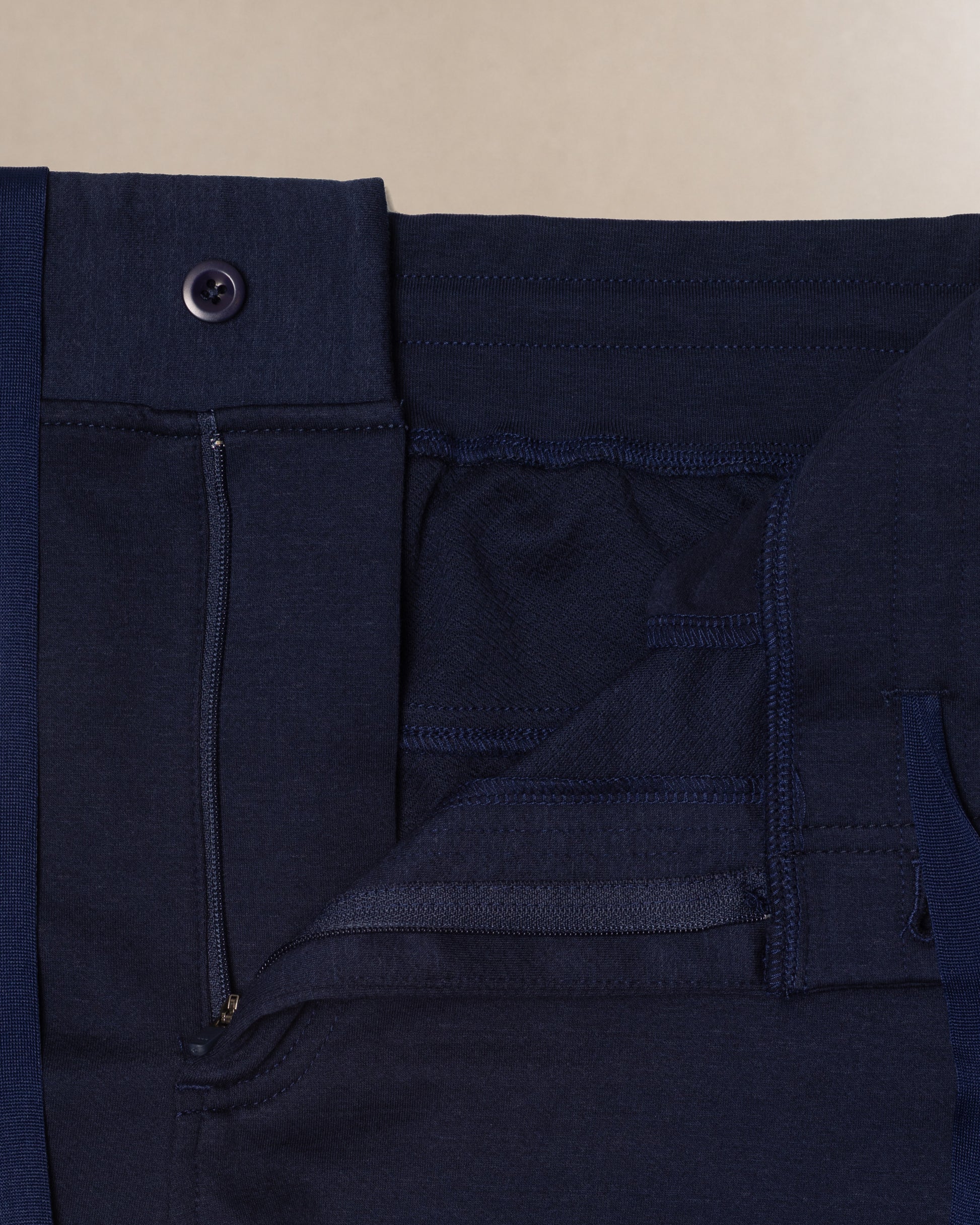 Close-up detail shot of dark navy blue scrub pants showing the waistband, button closure, belt loop, and front pocket stitching against a light beige background.