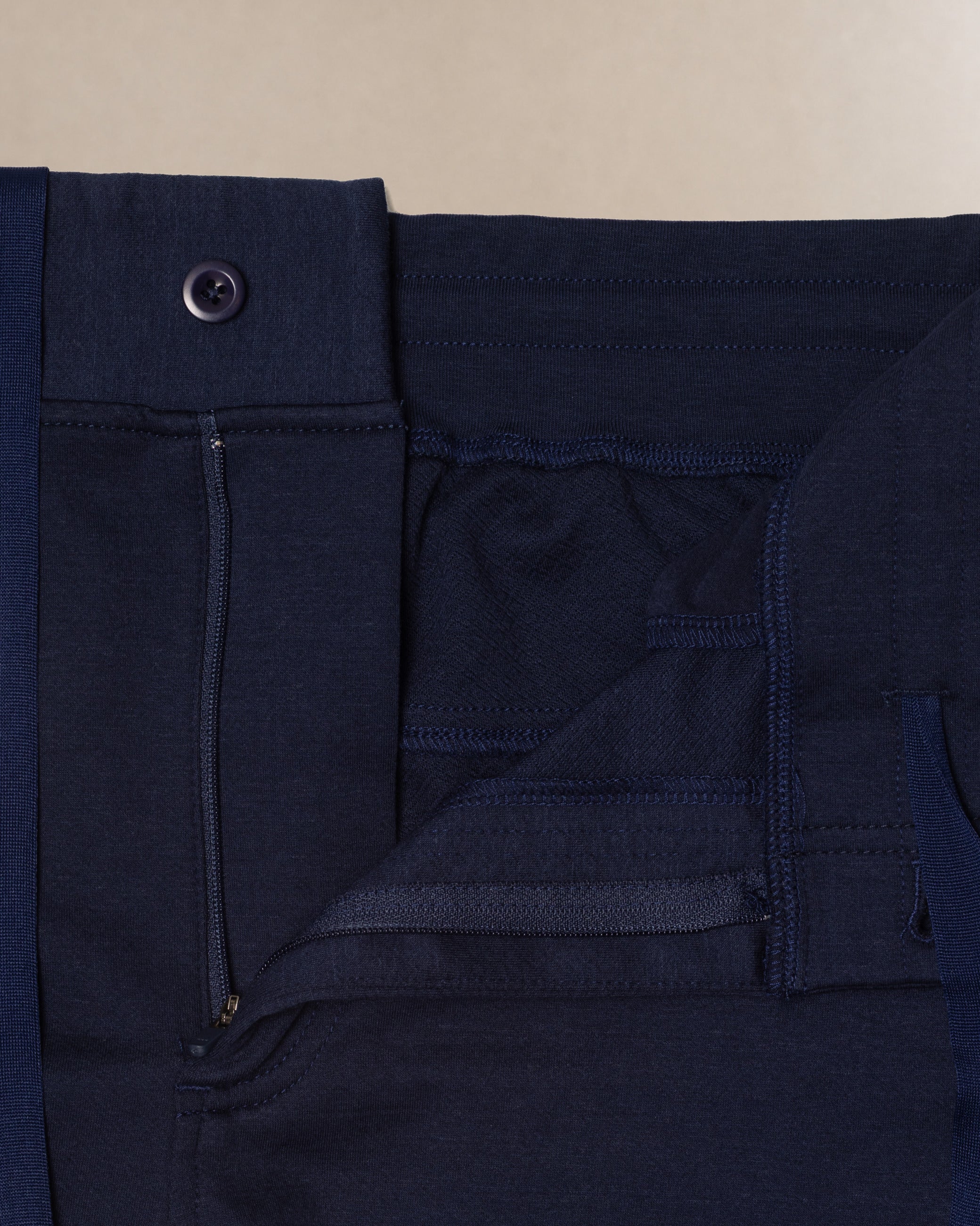 Close-up detail shot of dark navy blue scrub pants showing the waistband, button closure, belt loop, and front pocket stitching against a light beige background.