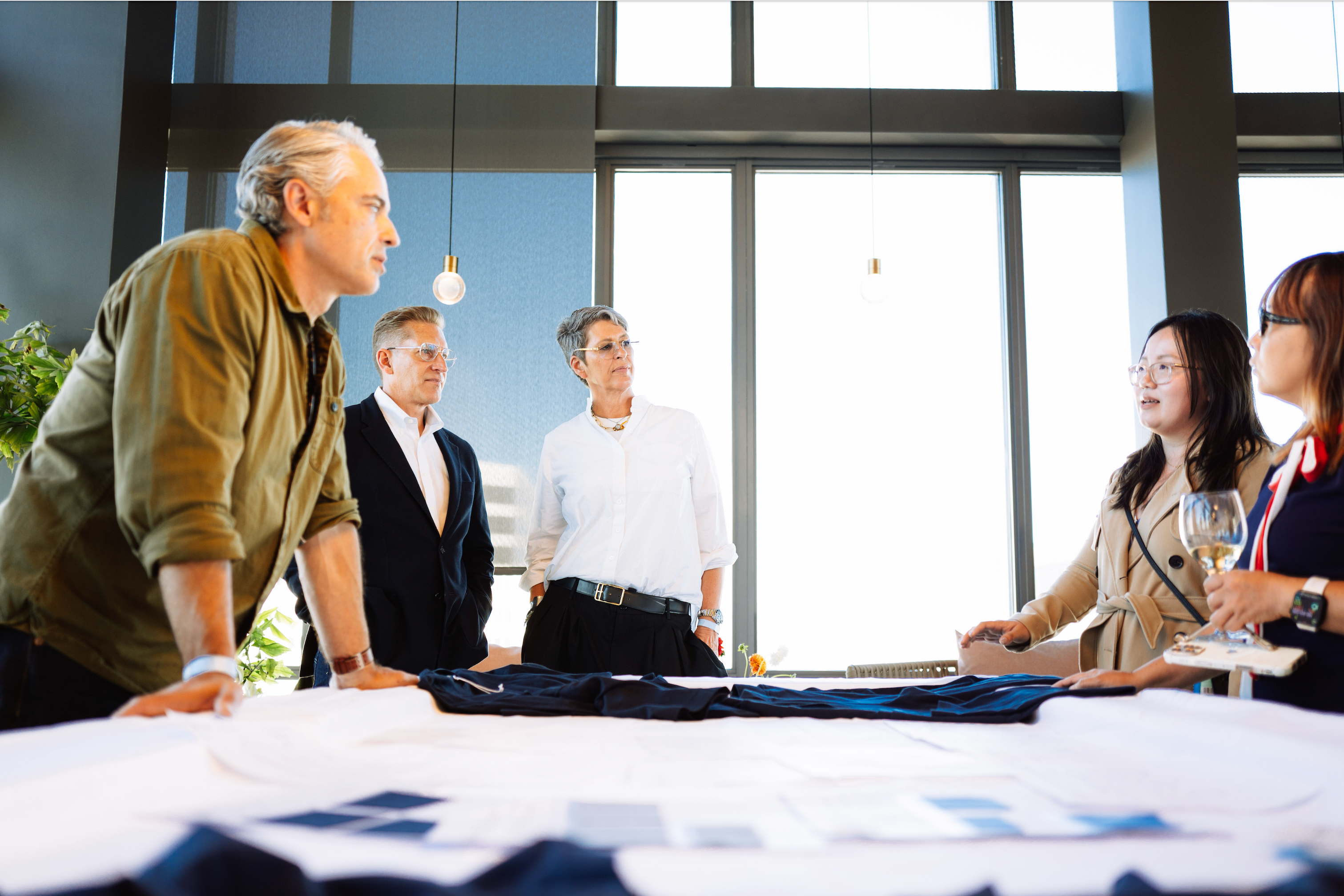 A group of five fashion industry professionals gathered around a table covered with fabric swatches and printed papers, with one man pointing at the materials. The room features large windows and modern architectural details.