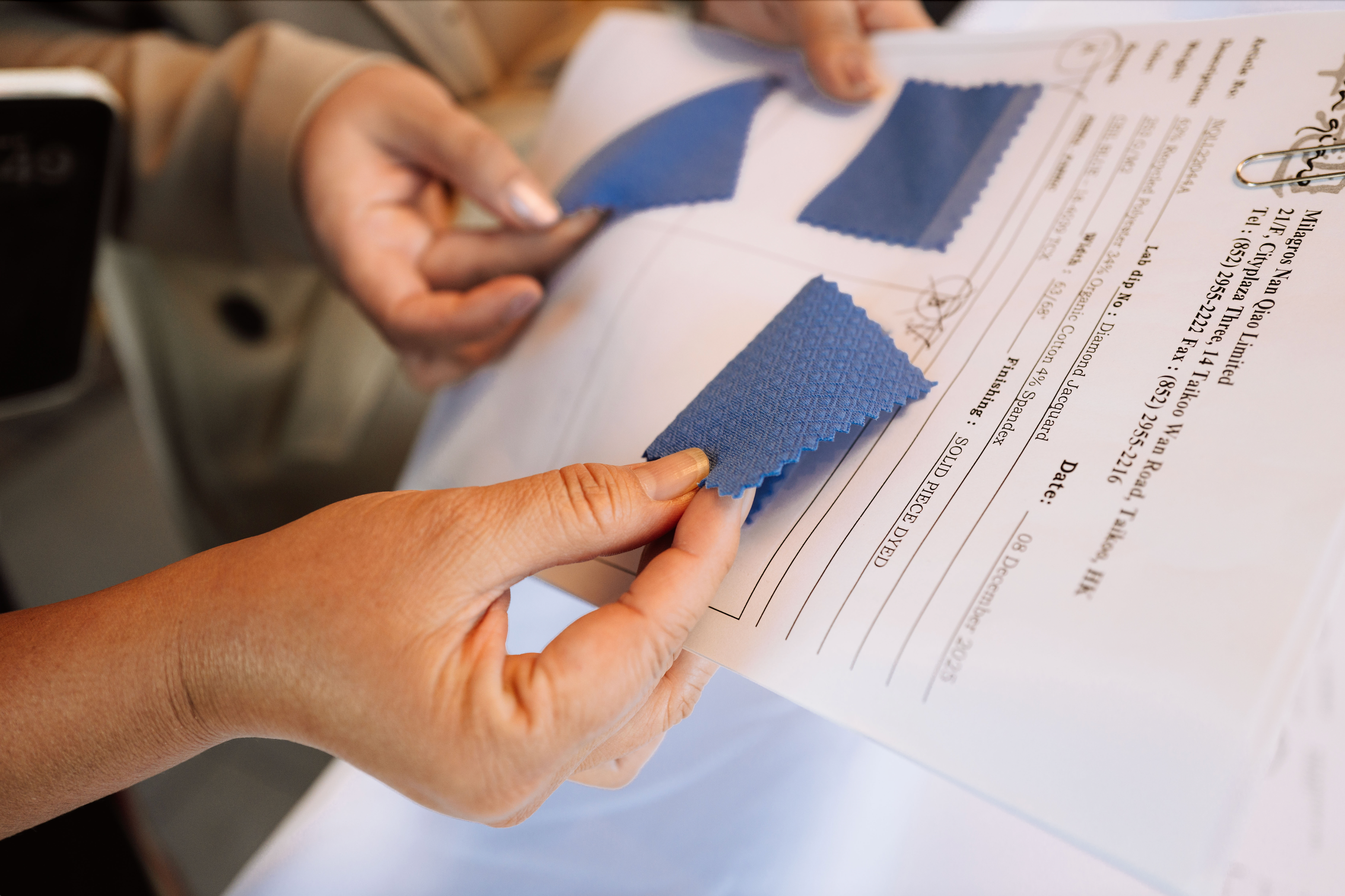A close-up of hands holding two textured blue fabric swatches against a white document, with a person in a beige blazer visible in the background.