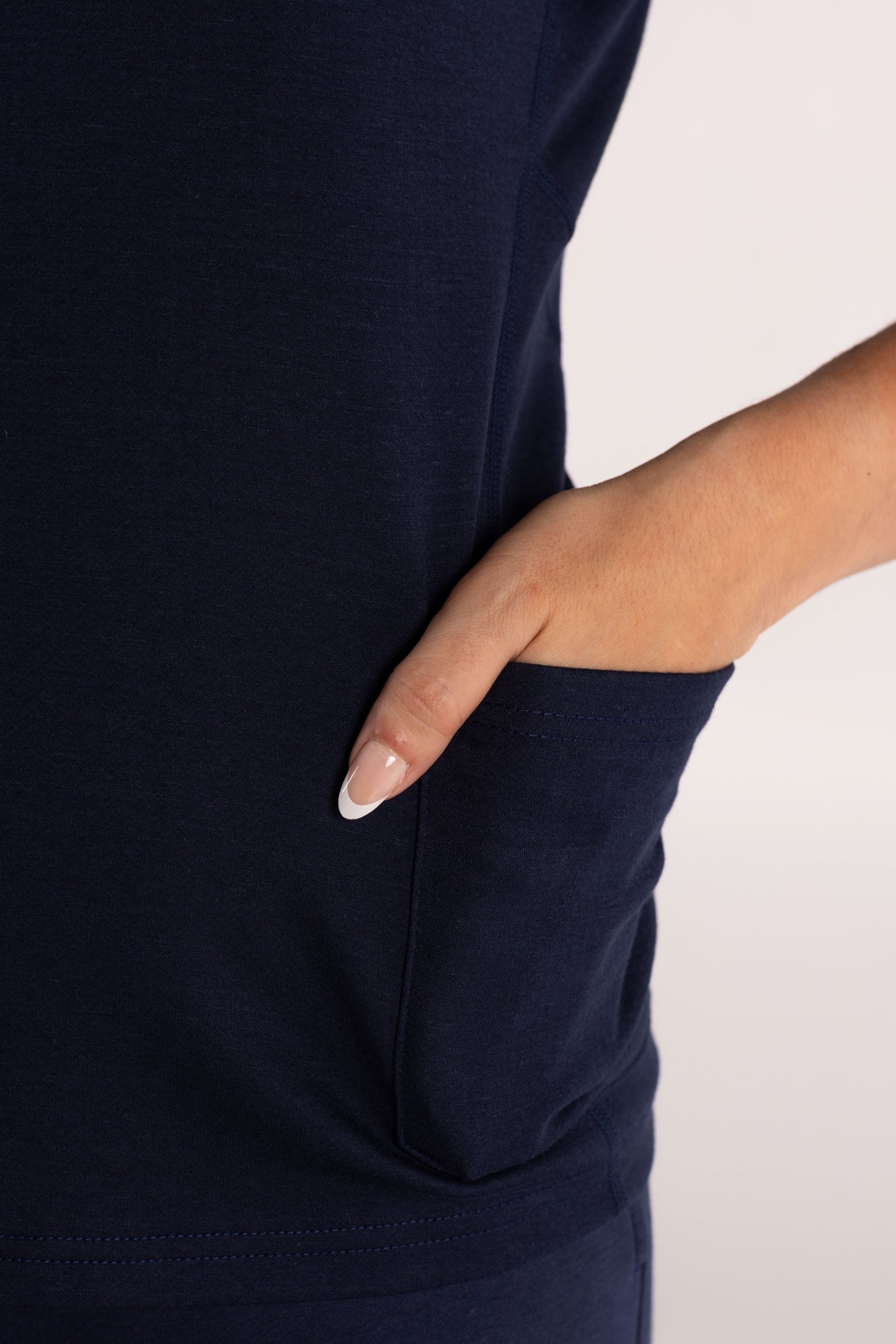 Close-up of a navy blue top with a side pocket, showing a hand with a white manicure sliding into the pocket against a light off-white background.
