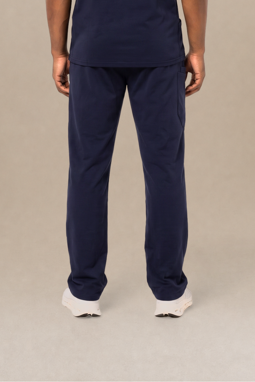 A man viewed from the back wearing a navy blue short-sleeve scrub shirt and matching navy blue scrub pants, standing against a neutral beige studio background.