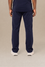 A man viewed from the back wearing a navy blue short-sleeve scrub shirt and matching navy blue scrub pants, standing against a neutral beige studio background.