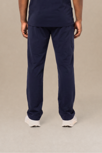 A man viewed from the back wearing a navy blue short-sleeve scrub shirt and matching navy blue scrub pants, standing against a neutral beige studio background.