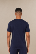 A man viewed from the back wearing a matching navy blue short-sleeve scrub shirt and straight-leg navy scrub pants, standing against a plain beige studio background. 
