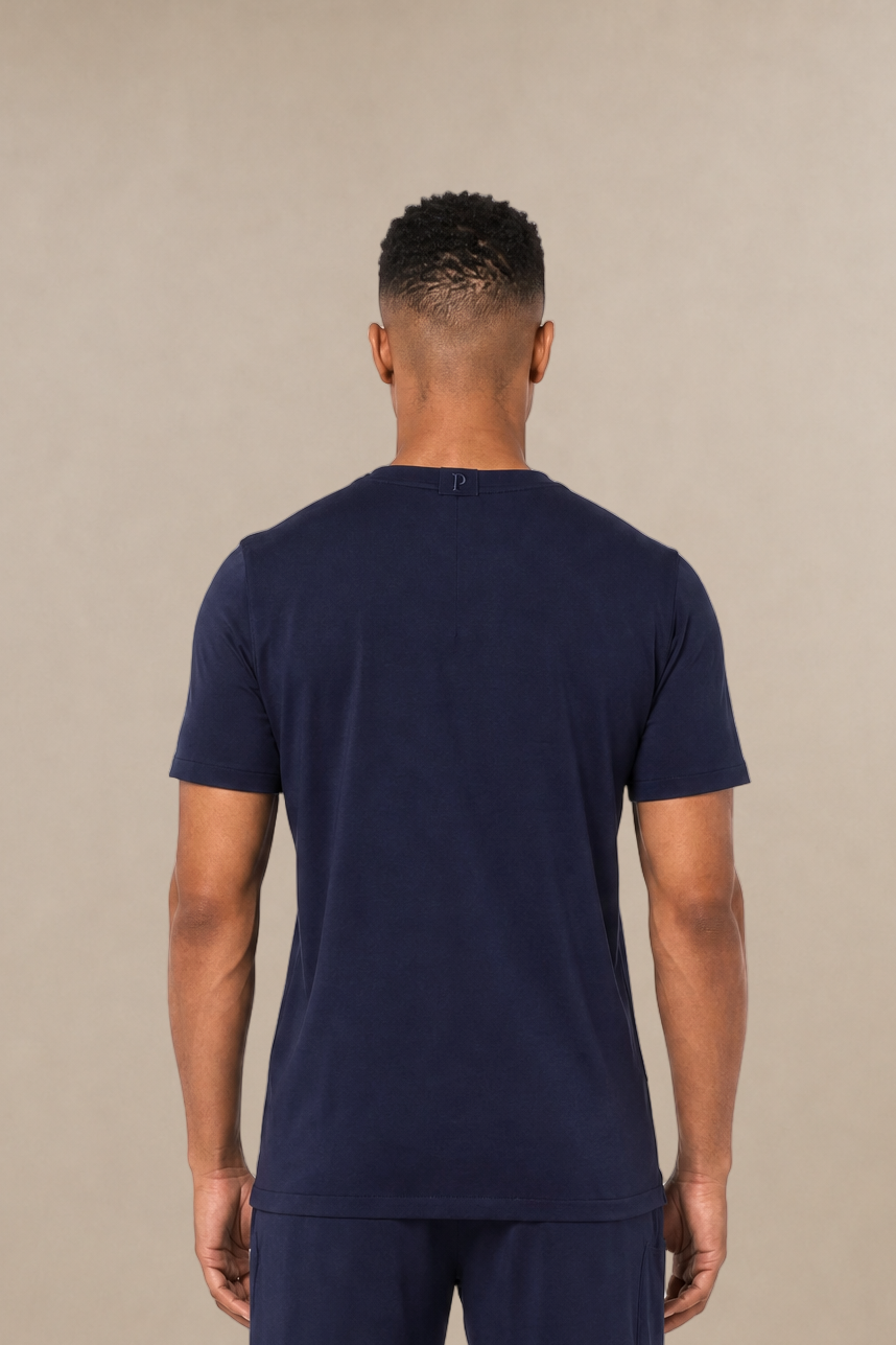 A man viewed from the back wearing a matching navy blue short-sleeve scrub shirt and straight-leg navy scrub pants, standing against a plain beige studio background. 