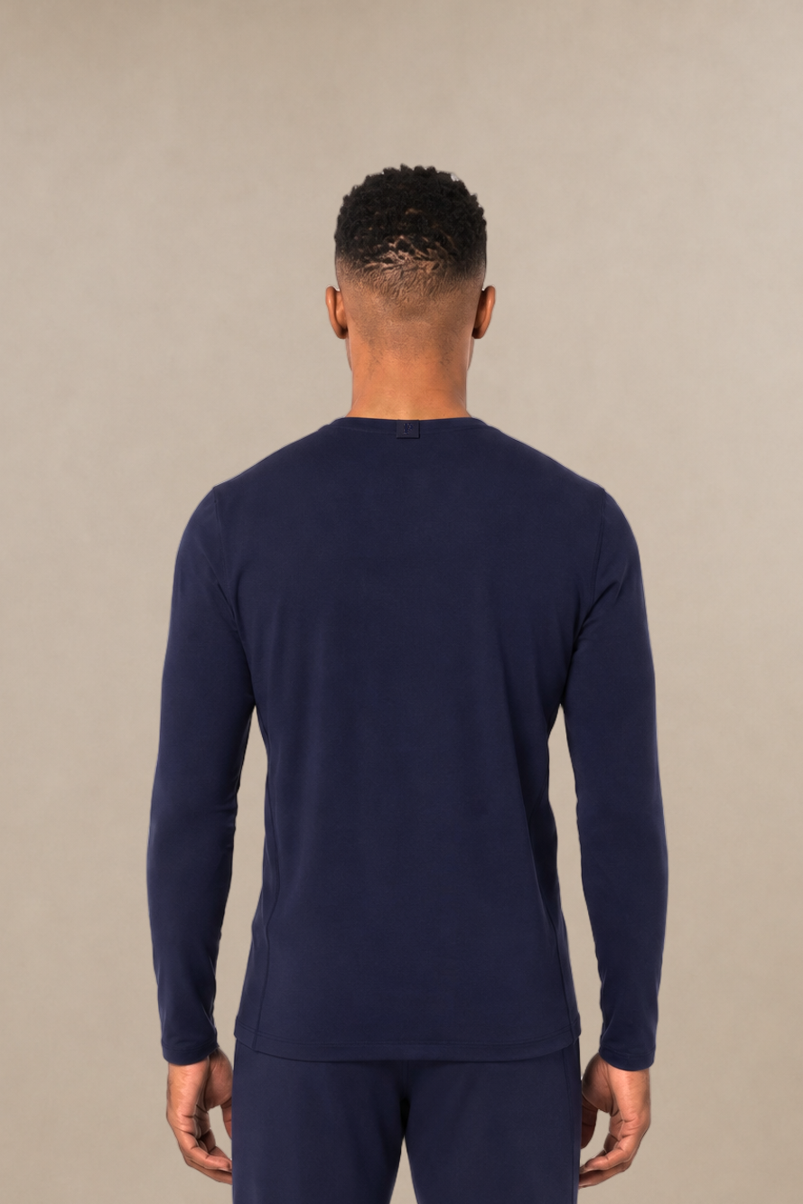 A man viewed from the back wearing a matching navy blue long-sleeve underscrub top and scrub pants, standing against a plain beige studio background.