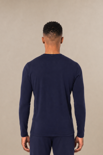 A man viewed from the back wearing a matching navy blue long-sleeve underscrub top and scrub pants, standing against a plain beige studio background.