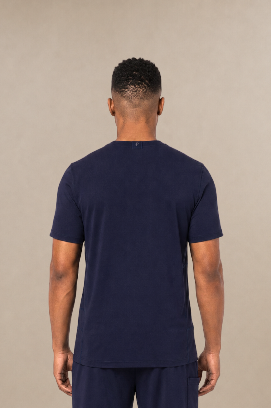 A man viewed from the back wearing a navy blue short-sleeve underscrub shirt and matching navy blue sweatpants, standing against a plain beige studio background.