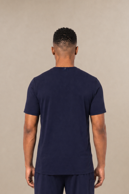 A man viewed from the back wearing a navy blue short-sleeve underscrub shirt and matching navy blue sweatpants, standing against a plain beige studio background.