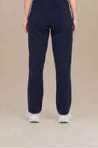 A woman viewed from the back wearing a navy blue scrub set consisting of a short-sleeve top and matching pants, standing against a solid beige studio background.