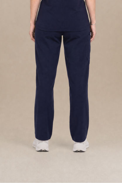 A woman viewed from the back wearing a navy blue scrub set consisting of a short-sleeve top and matching pants, standing against a solid beige studio background.