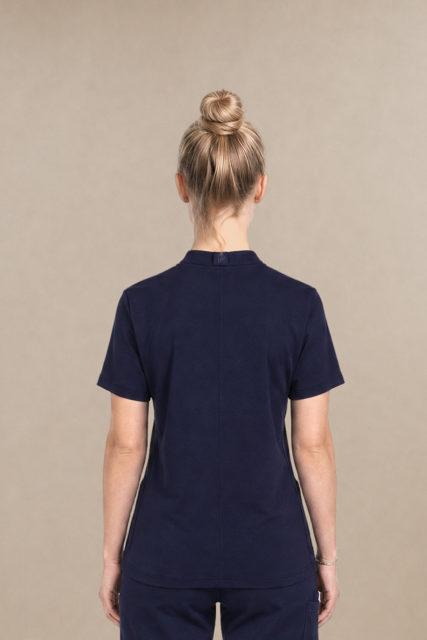 A woman viewed from the back wearing a navy blue scrub set consisting of a short-sleeve top and matching pants, standing against a solid beige studio background.