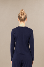 A woman standing with her back to the camera wearing a matching navy blue long-sleeve underscrub top and straight-leg scrub pants, against a plain beige studio background.