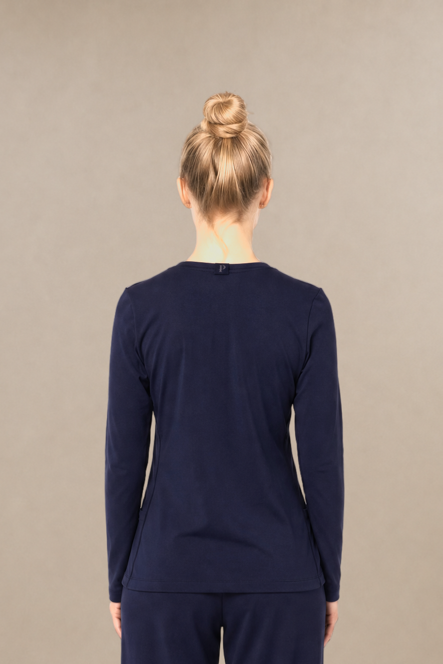 A woman standing with her back to the camera wearing a matching navy blue long-sleeve top and straight-leg pants scrub set, against a plain beige studio background.